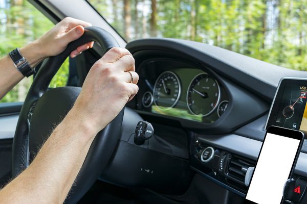Male hands holding car steering wheel. Hands on steering wheel of a car driving near the lake. Man driving a car inside cabin. Smartphone in holder with isolated white empty blank screen. Copy space